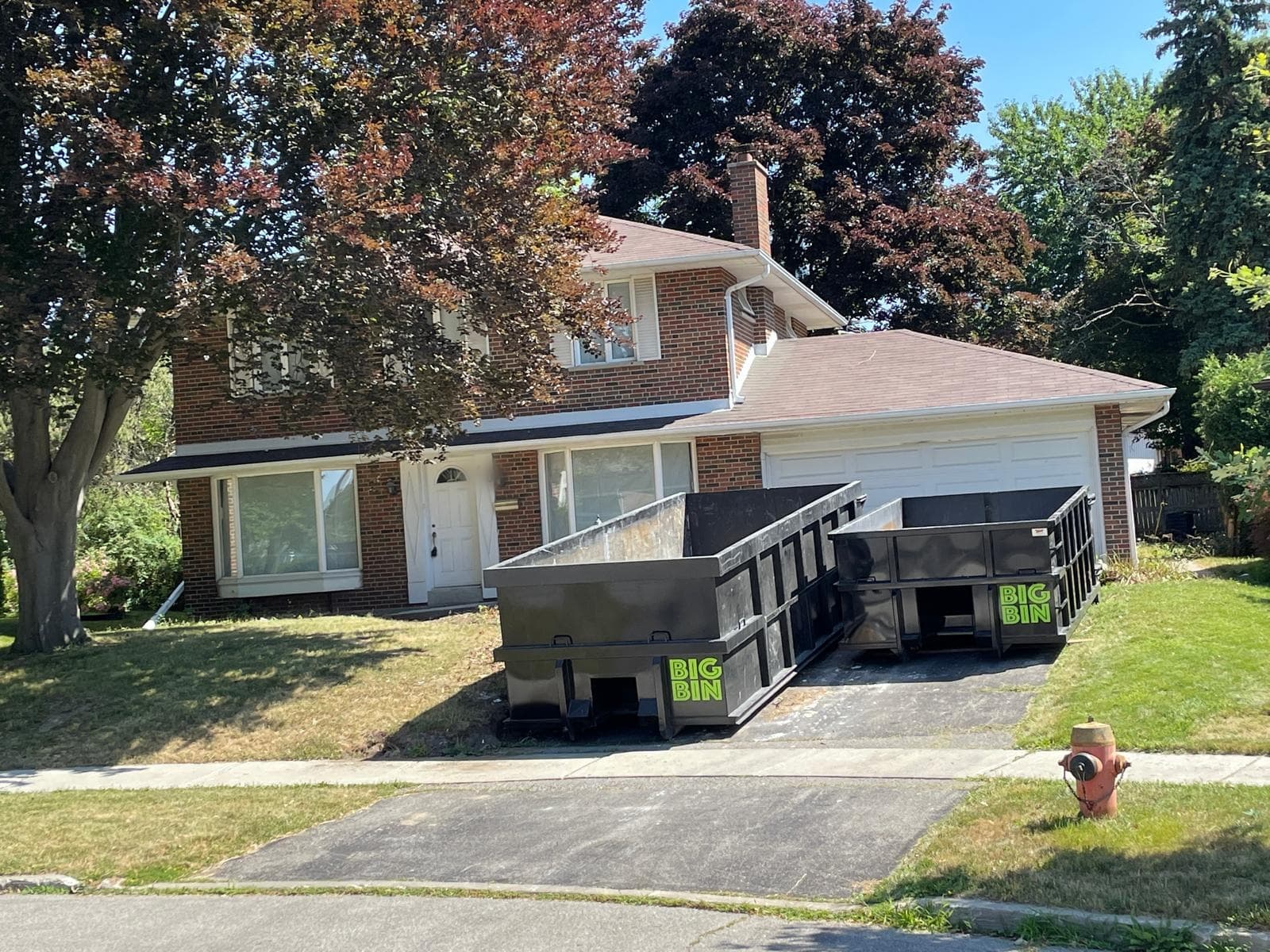 Two black Big Bin roll-off bins on a suburban brick-house driveway on a sunny day