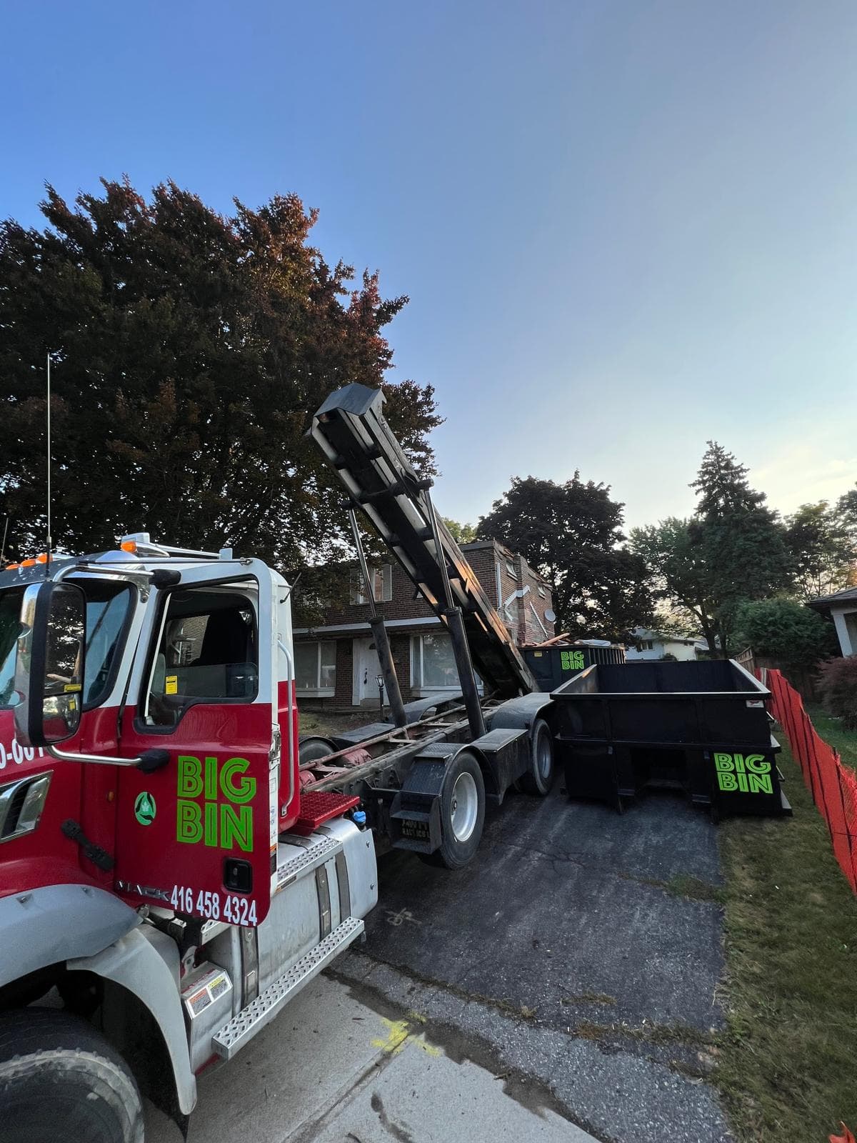 A Big Bin truck lowering a roll-off bin onto a residential driveway at golden hour